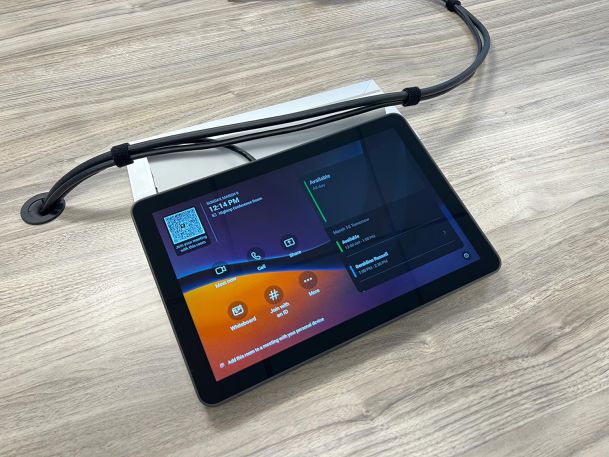 Touchscreen room scheduling panel sitting on wooden conference table surface with black wires