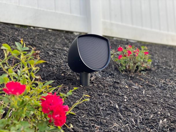 Outdoor ground speaker facing forward in a dark mulch landscape bed near red flower bushes