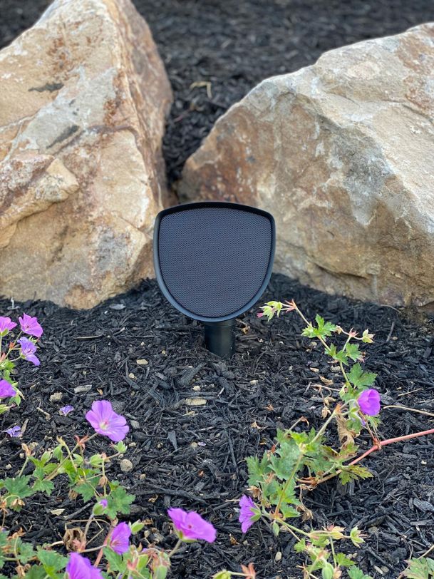 Black curved landscape speaker positioned on mulch ground between large natural boulders