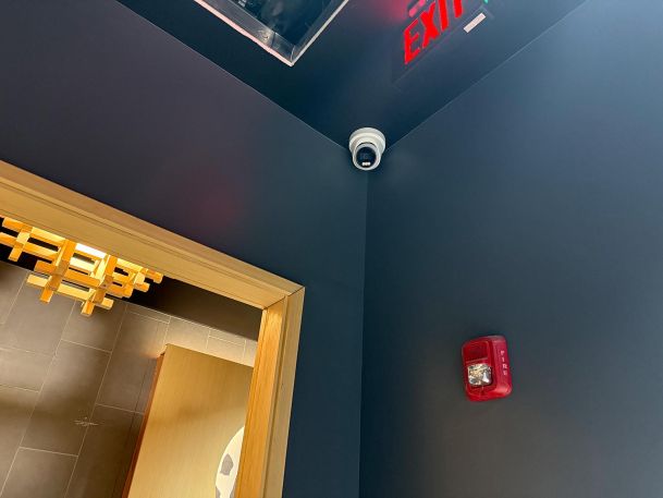 Corner ceiling security camera mounted above door frame near red fire alarm strobe