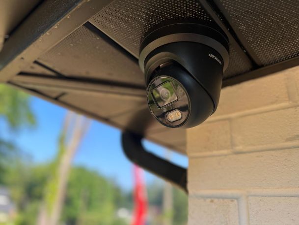 Black outdoor dome security camera mounted underneath a dark roof eave against brick wall