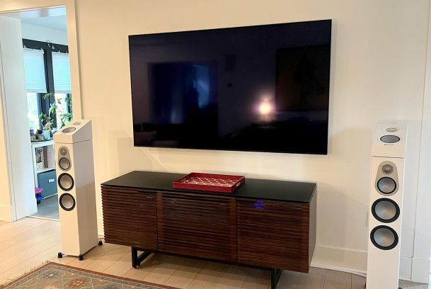 Flat screen television mounted above dark slatted cabinet flanked by white tower speakers