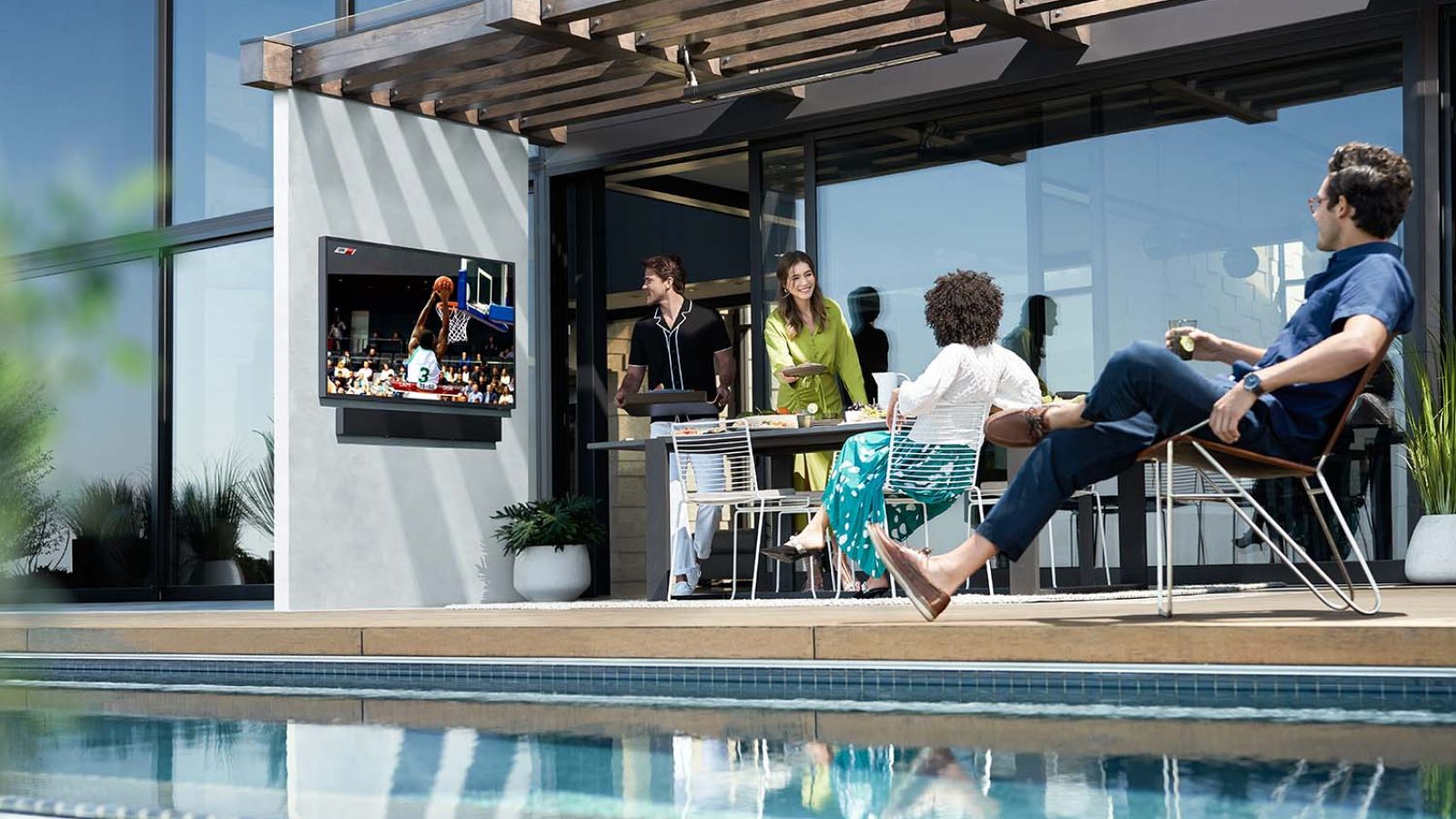 A group of people gathered in a landscaped backyard, enjoying a social gathering with outdoor seating and a TV displaying a basketball game.