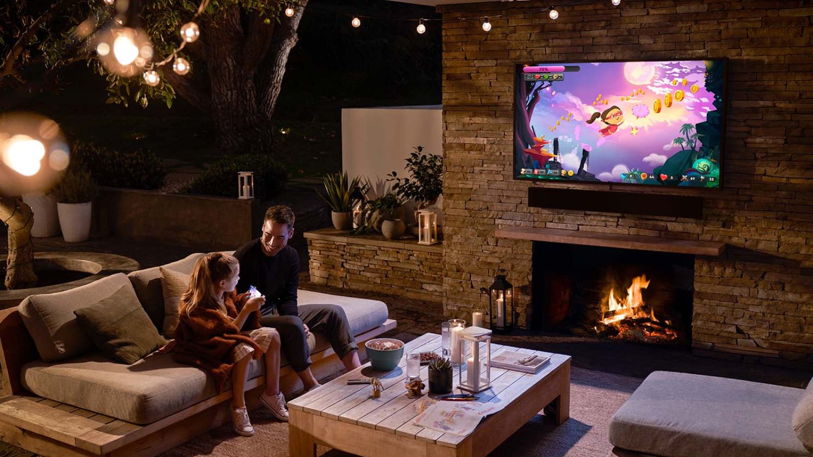 A father and daughter sitting on a cozy outdoor couch at night, enjoying quality time near a fireplace with string lights above, while a TV on the stone wall displays a colorful video game.